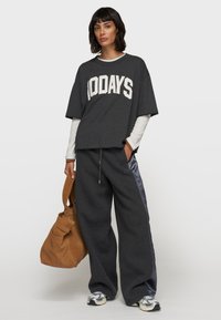 T-shirt noir oversized avec "100 JOURS" en lettres blanches, associé à un pantalon de jogging gris ample avec des panneaux en satin sur les côtés et un sac marron.