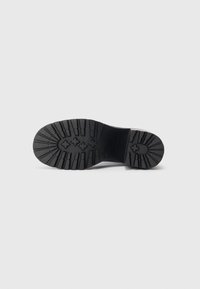 Semelle de botte noire face vers le haut avec de profonds motifs de semelle, incluant des formes croisées et en barres sur un fond blanc uni.