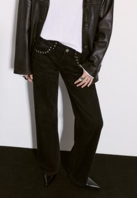 Čierne nohavice s širokými nohavicami a detailným zlatým nitovaním na vreckách, ktoré sú skombinované s bielym tričkom a čiernou koženou bundou, doplnené o lodičky so špicatou špičkou.