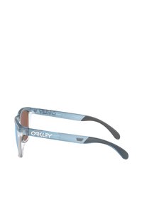 Occhiali da sole Oakley blu trasparenti con lenti marroni, caratterizzati da una presa in gomma nera texturizzata sulle tempie e logo bianco sulla montatura.