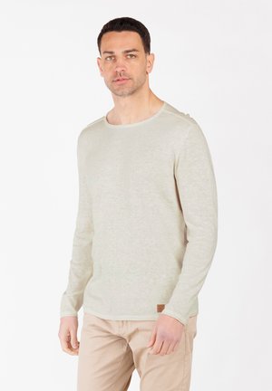Chemise beige clair à manches longues, fabriquée dans un tissu doux et texturé. Elle présente un col rond et un petit patch logo à l'ourlet.
