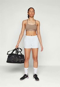 Καφέ αθλητικό σουτιέν και λευκά σορτς, στέκεται με μια μαύρη τσάντα γυμναστικής. Μαύρα παπούτσια με ροζ λεπτομέρειες και λευκές κάλτσες. Ουδέτερο φόντο.