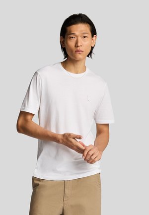T-shirt blanc à manches courtes en tissu doux, avec un col rond et un petit logo brodé sur la poitrine. Porté avec un pantalon beige.