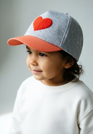 Young child wearing a gray and orange cap with a red heart, dressed in a white sweatshirt, looking slightly to the side and smiling softly.