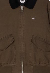 Veste zippée marron avec un col noir, dotée de deux grandes poches avant et d'un petit logo blanc "OBEY" sur la poitrine.