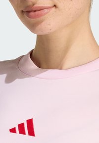 Jasnoróżowa koszulka z okrągłym dekoltem, z czerwonym logo w trzy paski po lewej stronie. Gładka faktura materiału.