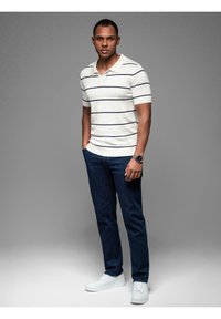 Krátký rukáv bílá polo tričko s námořnickými pruhy, zkombinované s tmavě modrými džíny a bílými teniskami. Model stojí vzpřímeně proti šedému pozadí.