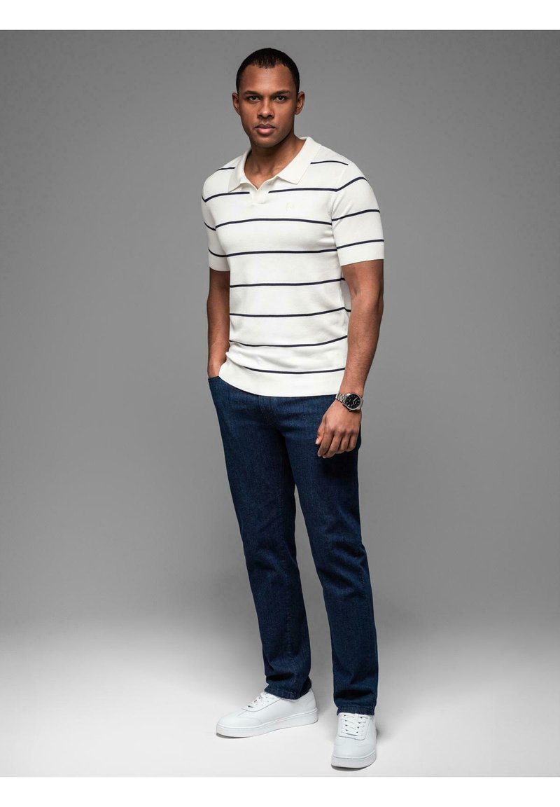 Krátký rukáv bílá polo tričko s námořnickými pruhy, zkombinované s tmavě modrými džíny a bílými teniskami. Model stojí vzpřímeně proti šedému pozadí.