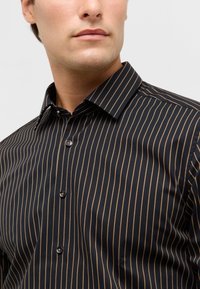 Zwarte shirt met dunne verticale bruine strepen, met knopen aan de voorkant, een kraag en een gladde textuur. Heeft zwarte knopen langs de voorkant.