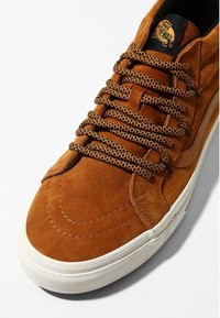 Brązowe zamszowe sneakersy z teksturowaną powierzchnią, czarnymi i brązowymi sznurowadłami w wzory, białą gumową podeszwą i logo Vans na języku.