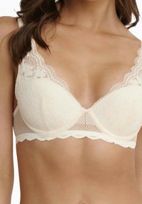 Lace-trimmed bra in cream with floral patterned cups, scalloped edges, and a central gold accent. Soft fabric with a smooth finish.
