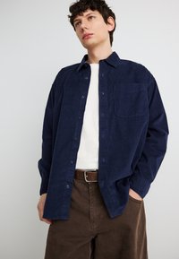 Jeune homme portant une chemise en velours côtelé bleu marine boutonnée sur un t-shirt blanc, avec un pantalon marron et une ceinture assortie, debout devant un fond uni.