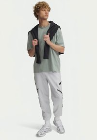 Jauns vīrietis ar cirtainiem matiem, ģērbies gaiši zaļā lielizmēra T-kreklā, baltās biksēs ar melniem akcentiem, baltos sporta apavos un melnā džemperī, kas uzvilkts pār pleciem.