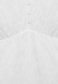 Robe blanche en tissu texturé ressemblant à de la dentelle. Le corselet a un col rond et six petits boutons sur le devant.