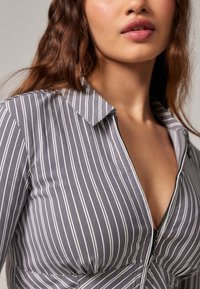 Camisa de rayas gris y blanca con cuello, que presenta un cierre frontal y una cintura ajustada. La tela parece suave con un ligero brillo.