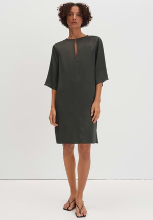 Vrouw met kort krullend haar, draagt een losse, donkergroene knielange jurk met ellebooglange mouwen en zwarte sandaaltjes met bandjes.