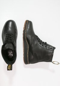Un par de botas negras de cuero con cordones y suelas marrones, una mostrada desde arriba y la otra de lado sobre un fondo blanco.