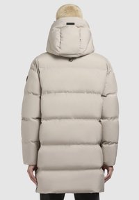 Chaqueta de plumas beige con una gran capucha, diseño acolchado y puños elásticos. Presenta una textura suave y mate, con un logo mínimo en el cuello.