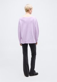 Άτομο με κοντά ξανθά μαλλιά που φοράει φαρδιά λιλά πουλόβερ, μαύρο παντελόνι καμπάνα και μαύρα παπούτσια, στέκεται με την πλάτη προς το φακό σε λευκό φόντο.