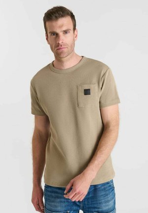Khakifarbenes T-Shirt mit kurzen Ärmeln aus strukturiertem Stoff und einer Brusttasche mit einem quadratischen schwarzen Akzent. Getragen mit blauen Jeans.