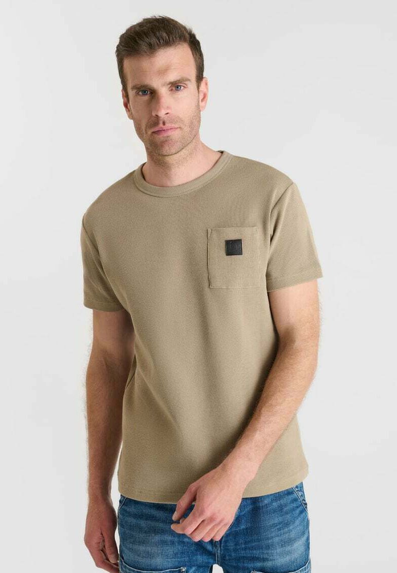 Camiseta de manga corta color caqui con una tela texturizada y un bolsillo en el pecho que presenta un detalle cuadrado negro. Llevada con jeans azules.