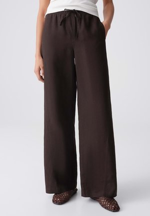 Frau trägt dunkelbraune, weit geschnittene Hosen mit Kordelzugbund, ein weißes ärmelloses Oberteil und gewebte dunkelbraune Slipper.