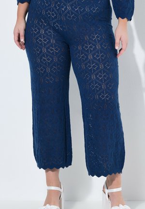 Marineblauwe gehaakte broek met geometrische diamantpatronen, gedragen met witte sandalen met hoge hakken.