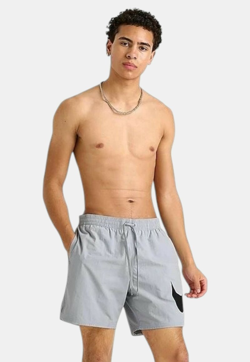 Pantaloni da bagno grigi con cintura elastica, cordino e un dettaglio del logo nero sul lato sinistro. Tessuto leggero e liscia.