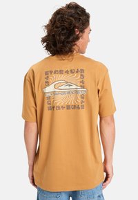 Camiseta de manga corta color ámbar hecha de algodón. Presenta un gran estampado gráfico en la parte posterior con líneas curvas y texto en colores contrastantes.