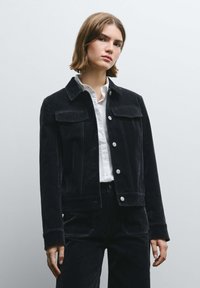 Giacca in velluto nero a coste con due tasche sul petto, bottoni argento e cuciture a contrasto, indossata sopra una camicia bianca con bottoni.