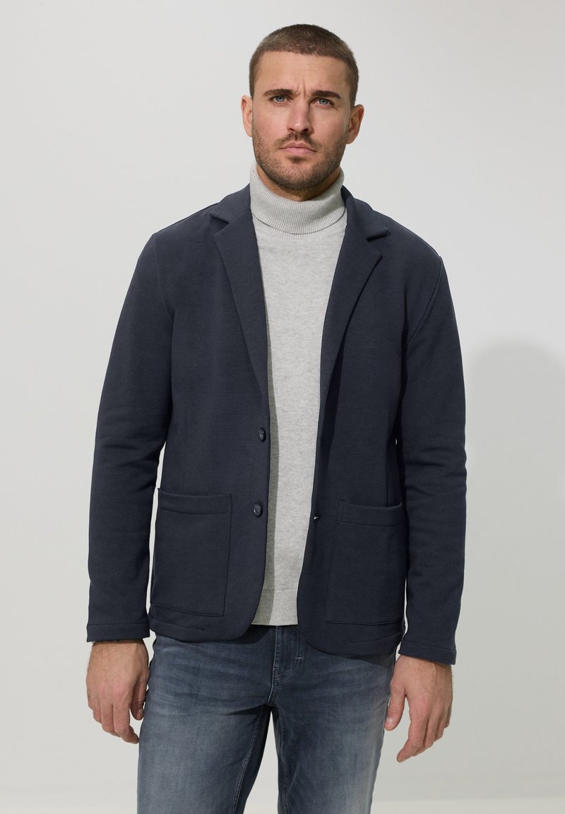 Man met kort haar, gekleed in een donkerblauw colbert over een lichtgrijze coltrui en blauwe spijkerbroek, staand tegen een effen witte achtergrond.