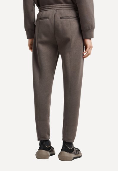 Persona che indossa pantaloni jogger grigi aderenti con tasche posteriori a filetto e sneaker nere con suole spesse color beige, in piedi di spalle.