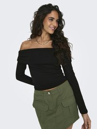 Žena sa usmieva, má na sebe čierny top s dlhými rukávmi spustenými z plecí a olivovo zelenú sukňu s veľkými prednými vreckami, stojaca pred jednoduchým pozadím.