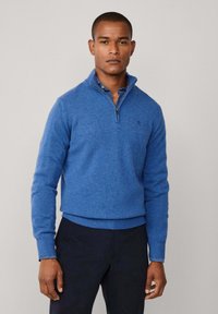 Suéter azul con cremallera en el cuello hecho de una tela texturizada, con un dobladillo y puños acanalados, y un sutil detalle del logo en el pecho.