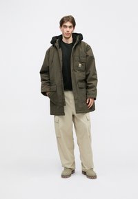 Parka verde con cappuccio e foderatura in pelliccia sintetica nera, con tasche frontali; abbinata a pantaloni cargo beige e stivali grigi.
