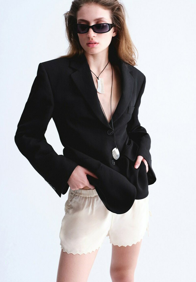 Femme portant un blazer noir, un short crème à bords ondulés, des lunettes de soleil noires et deux longs colliers avec pendentifs, posant les mains dans les poches.