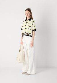 Short-sleeve polo shirt in textured fabric with a light green and brown abstract pattern, black collar, paired with wide white trousers and beige shoes.