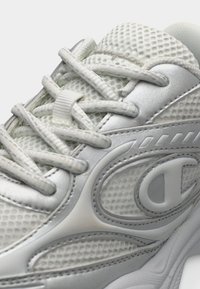 Λευκό αθλητικό παπούτσι με πλεκτό επάνω μέρος και ασημί λεπτομέρειες, υφασμάτινες επιφάνειες και επίπεδα κορδόνια. Η σχεδίαση περιλαμβάνει ένα εξέχον λογότυπο.