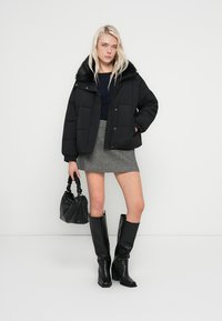 Crna podstavljena jakna s ovratnikom od umjetnog krzna, siva suknja, crne čizme do koljena i mala crna torbica. Glatke teksture i suptilni detalji.