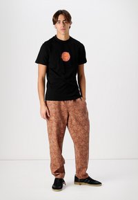 Black cotton t-shirt with circular orange graphic, paired with brown patterned pants, featuring abstract designs and black footwear.