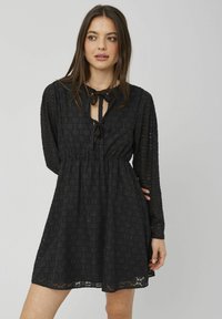Robe noire à manches longues, en tissu texturé avec un motif de cercles, et un décolleté en forme de clé lié par un nœud. Détail à taille élastique.