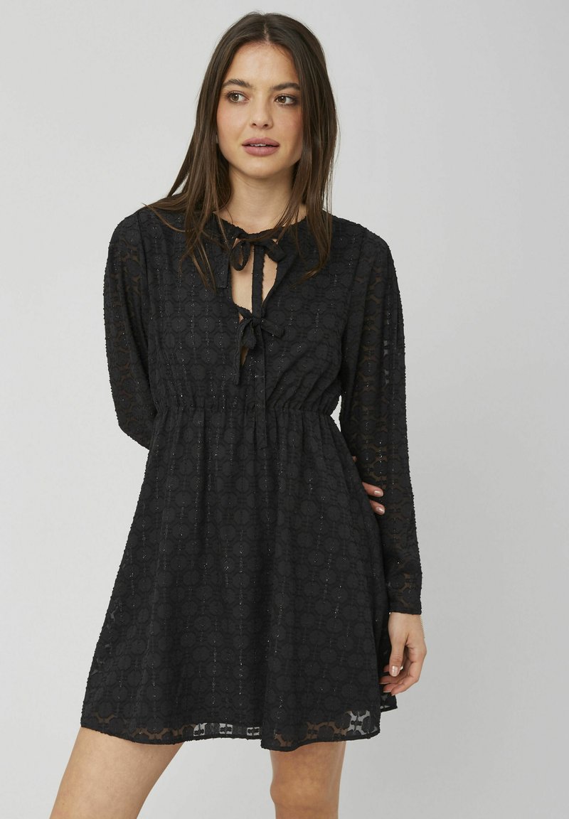 Robe noire à manches longues, en tissu texturé avec un motif de cercles, et un décolleté en forme de clé lié par un nœud. Détail à taille élastique.