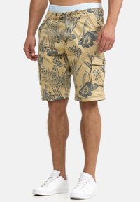 Shorts cargo beiges avec des motifs floraux sombres, plusieurs poches et une ceinture portant le nom « INDICODE » en bleu clair. Des sneakers blanches sont portées.
