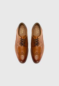 Zapatos de vestir de cuero marrón con detalles de brogue, que presentan una puntera redondeada, diseño con cordones y una textura suave. El color de la suela contrasta con la parte superior.