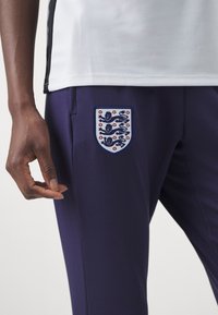 Pantalones atléticos morados con un escudo bordado de Inglaterra en el lado izquierdo. Hechos de una tela suave y elástica con un diseño ajustado.