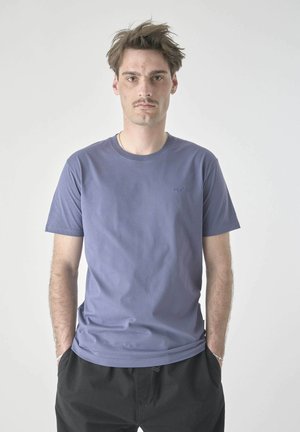 Junger Mann mit kurzem braunem Haar und Schnurrbart, der ein schlichtes blaues T-Shirt und schwarze Hosen trägt, steht mit den Händen in den Taschen vor weißem Hintergrund.