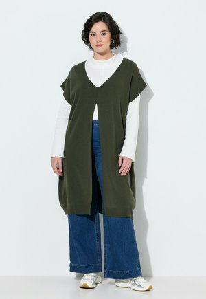 Kvinde med kort mørkt hår iført en hvid langærmet top, olivengrøn ærmeløs vest, blå jeans med vide ben og hvide sneakers stående mod en hvid væg.