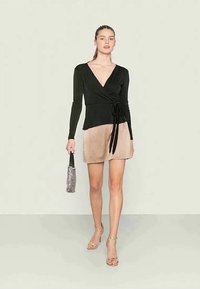 Mulher a caminhar, vestindo uma blusa preta envolvente, saia mini de seda bege, segurando uma pequena mala estampada e sandálias de salto douradas.