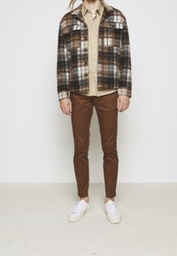 Brun rutig ulljacka över en ljusbeige skjorta, ihop med bruna slim-fit-byxor och vita sneakers med texturerade sulor.