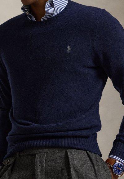 Marineblauwe trui over gestreept blauw-wit overhemd, grijze geplooide broek en blauwe horloge met bruine leren band om de pols.